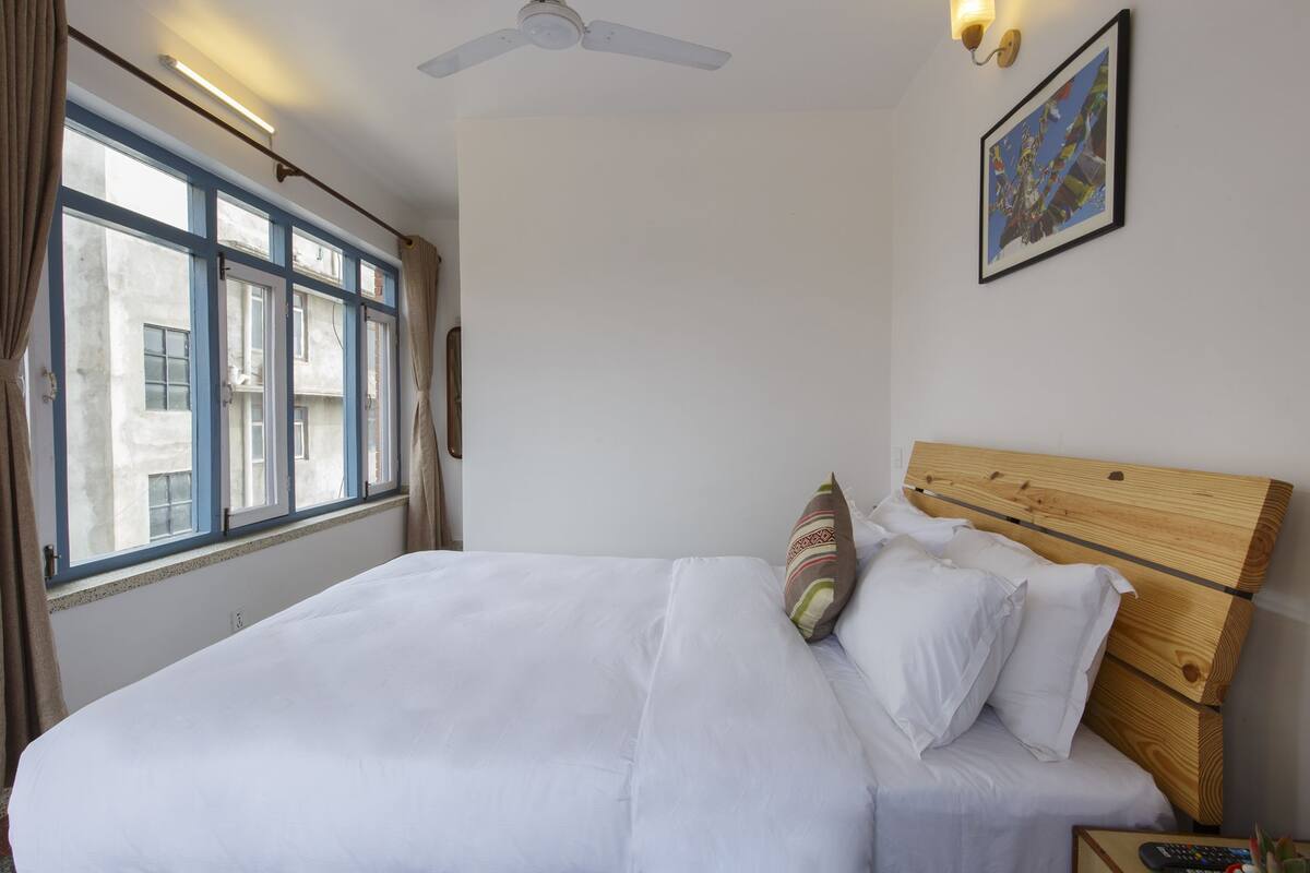 A neatly made bed with white linens and a wooden headboard, a ceiling fan, and a window with curtains.