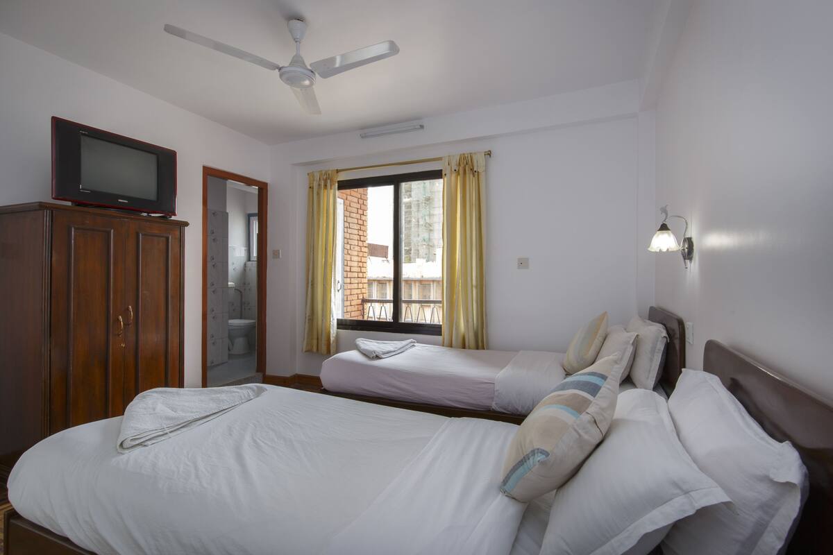 A hotel room with two single beds, a wooden wardrobe, a television on top, a ceiling fan, a window with curtains, and a wall-mounted light fixture.