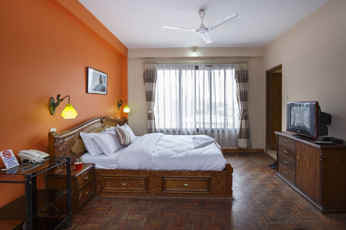 A hotel room with a bed, wooden headboard, a desk with a telephone, a television on a cabinet, and a window with curtains.