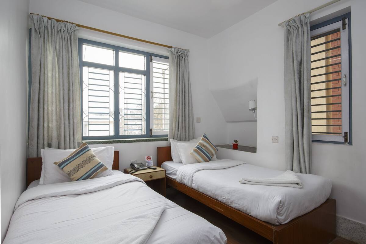A twin room with two single beds, a small bedside table with a phone, and a window with blinds.