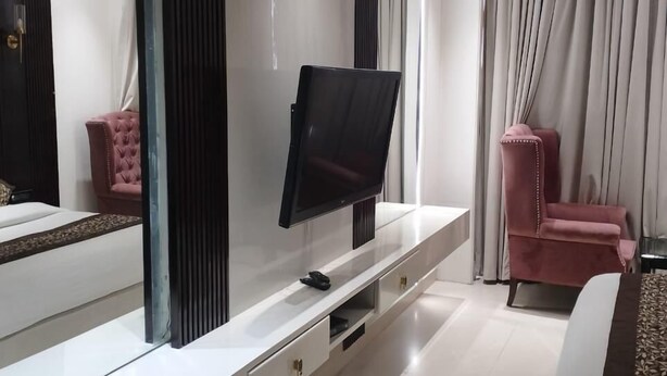 A modern hotel room with a large flat-screen TV on a white console, a pink chair, and a bed with a patterned bedspread.