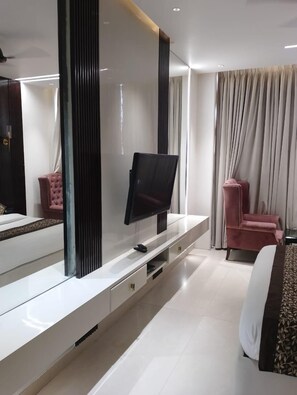 Habitación de hotel moderna con un televisor de pantalla plana grande sobre una consola blanca, una silla rosa y una cama con colcha estampada.
