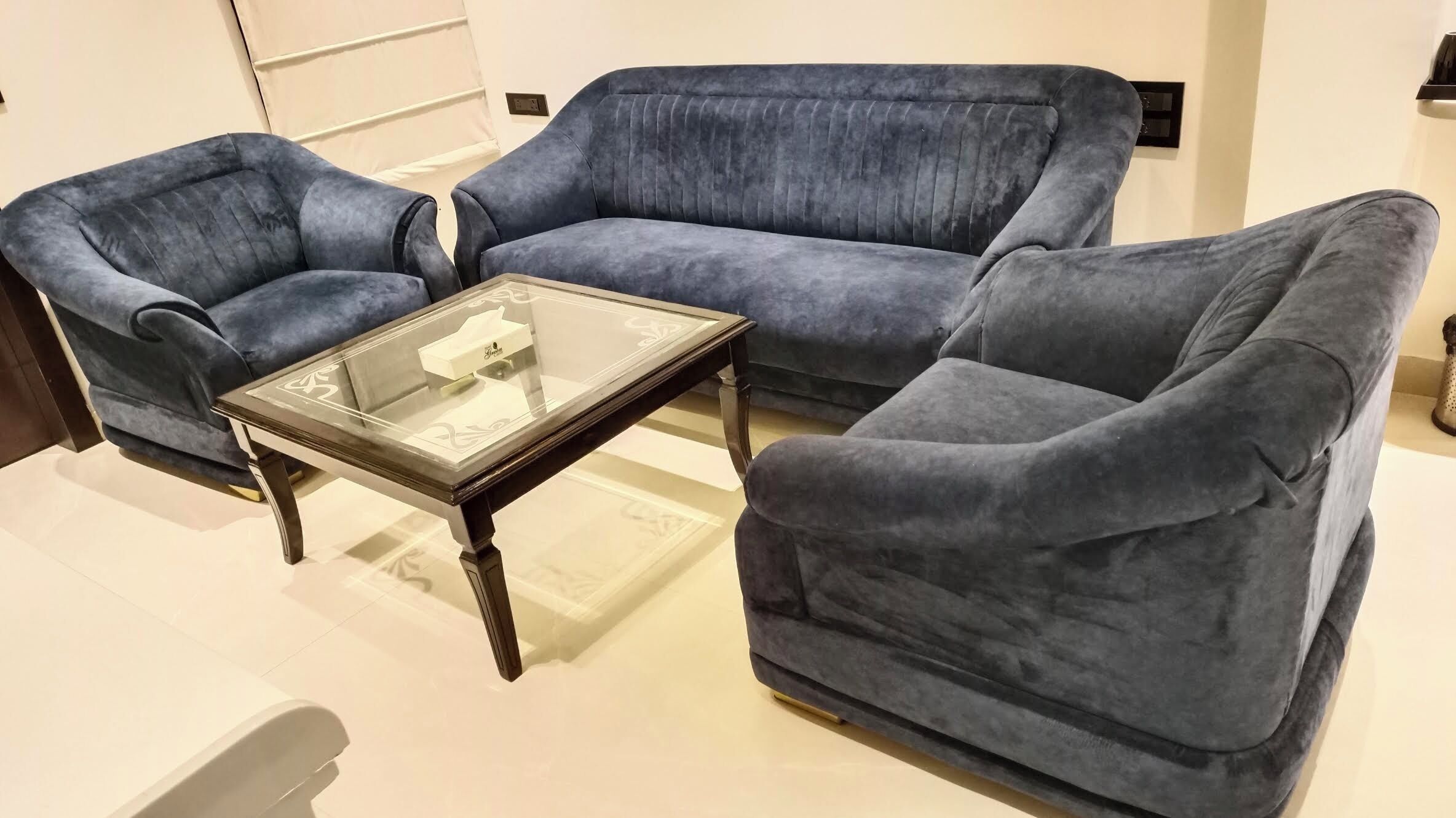 A room with a blue sofa set, a glass coffee table, and a white chair.