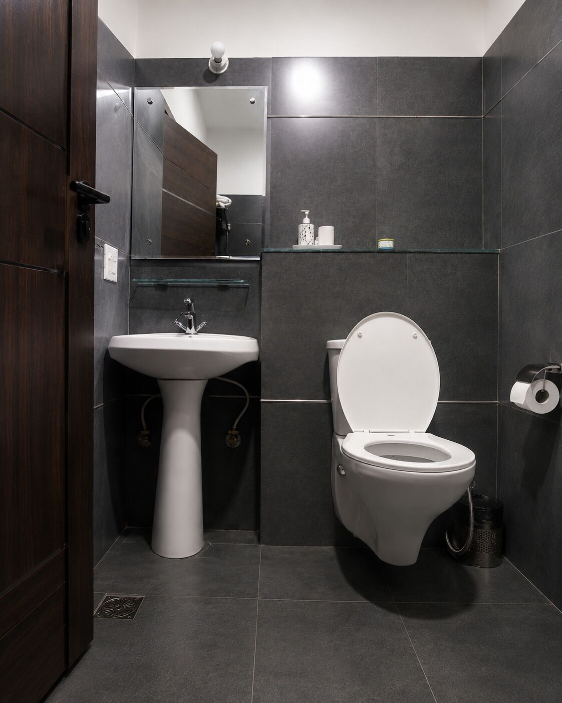 A modern bathroom with a white pedestal sink, a toilet, and a glass shelf with toiletries.