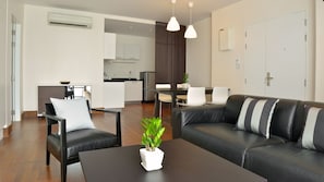 Ein modernes Wohnzimmer mit einer schwarzen Ledersofa, einem schwarzen Couchtisch und einer weißen Topfpflanze. Es gibt eine Essecke mit weißen Stühlen und eine kleine Küche im Hintergrund.