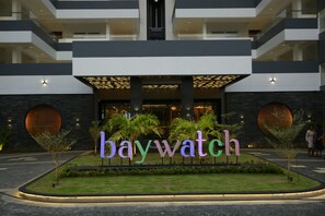 Entrada de hotel com um letreiro 'baywatch', design moderno e área ajardinada com palmeiras.