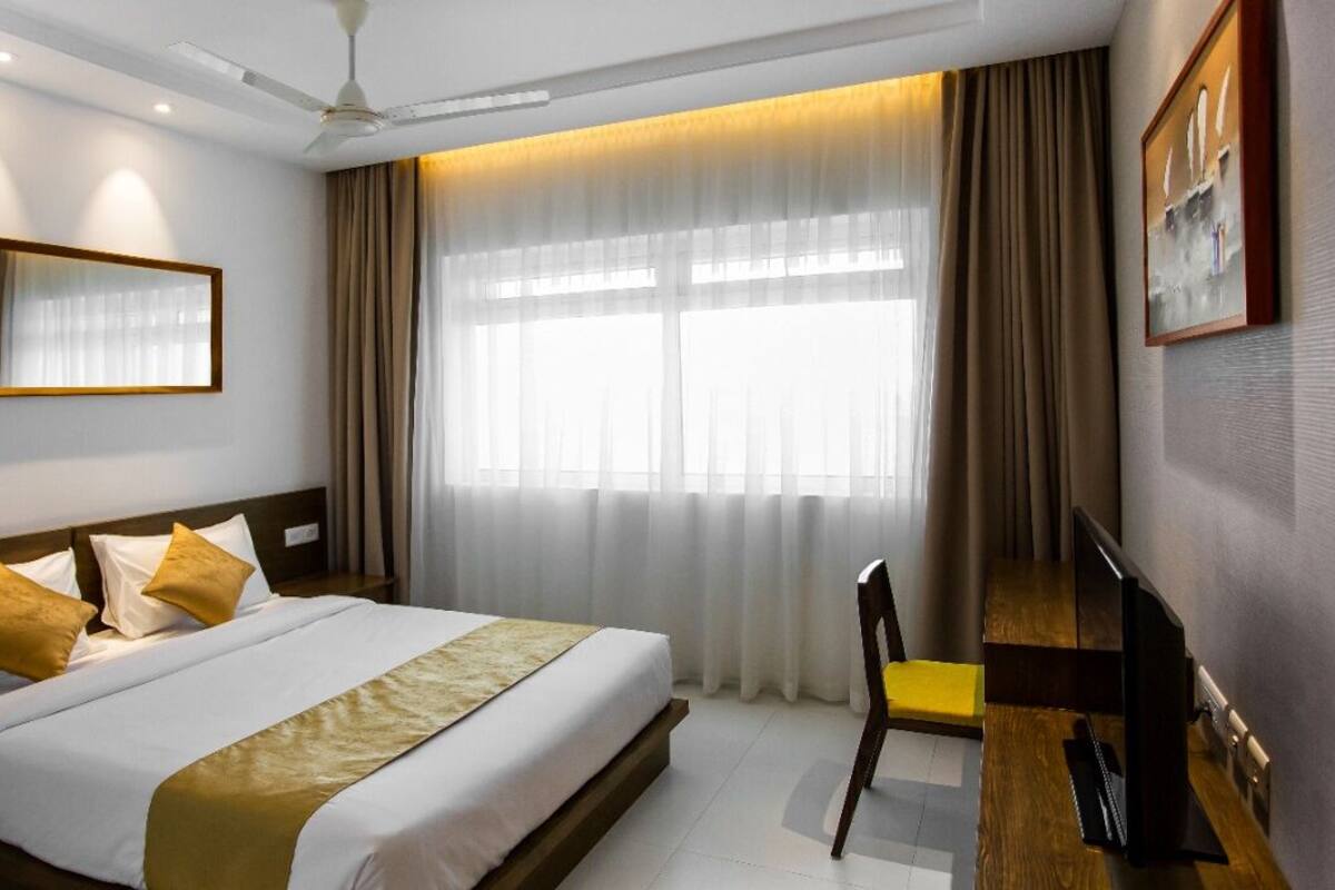 Una habitación de hotel con una cama, un escritorio, una silla, un televisor, un espejo y un gran ventanal con cortinas.