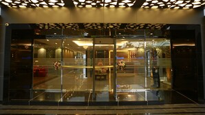 A hotel entrance with a glass facade, a geometric ceiling design, and a reflection of the interior. - Kent Baywatch Suites (Kochi)