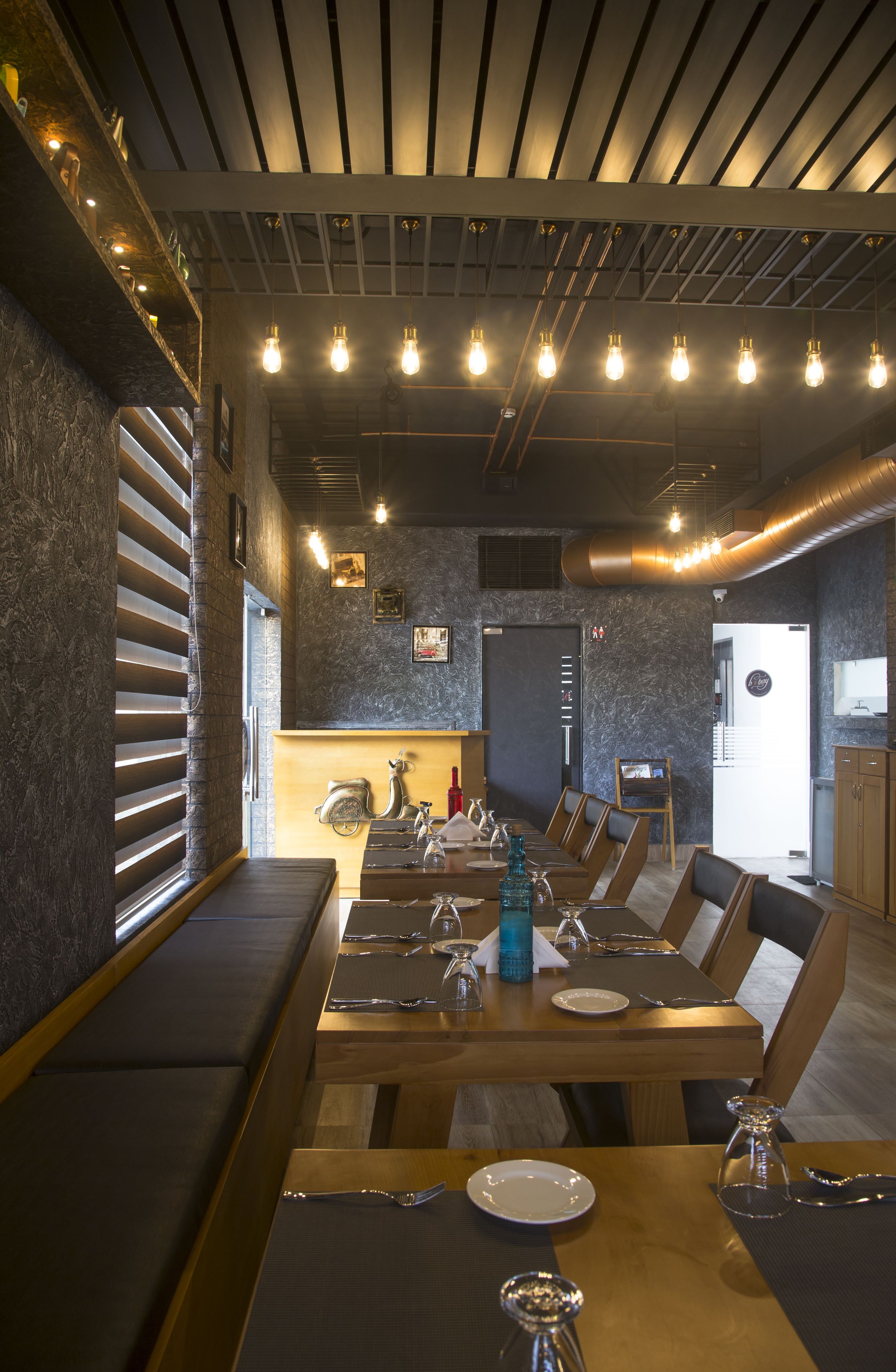 Restaurant amb una taula de fusta llarga preparada per dinar, zona de seients amb un banc i un disseny de sostre modern amb bigues i il·luminació a la vista.