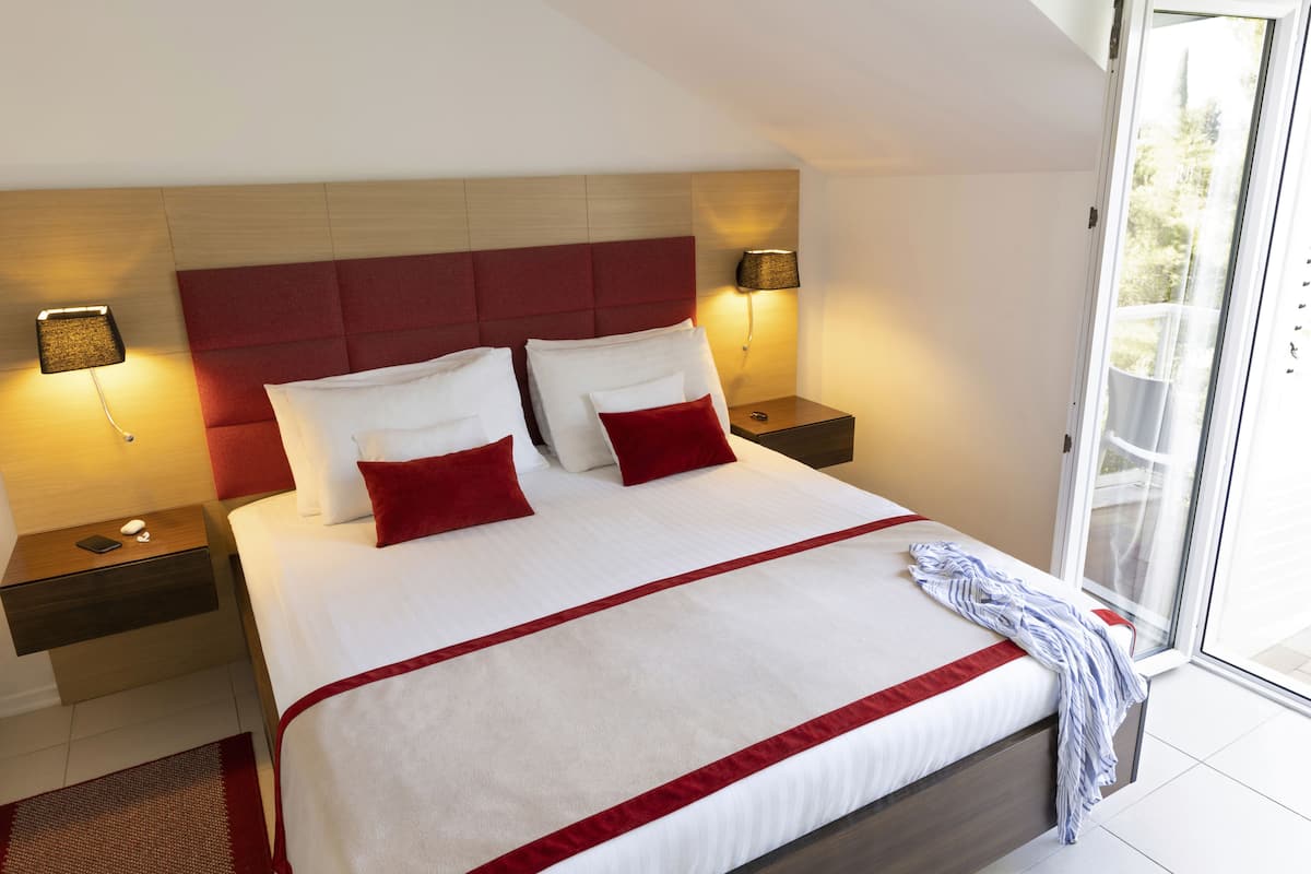 A hotel room with a large bed, red and white bedding, two bedside tables with lamps, and a view through a large window.