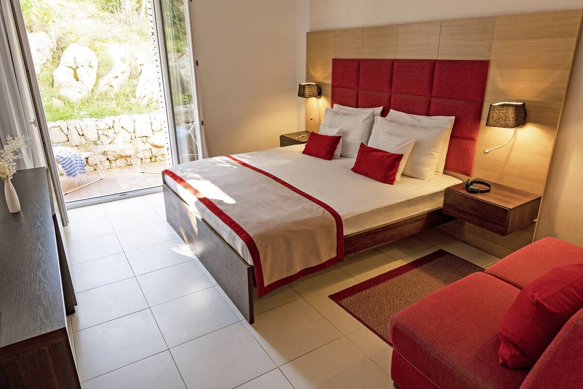 A modern hotel room with a large bed, red and white bedding, two bedside lamps, a red sofa, and a view of the outdoors through a glass door.
