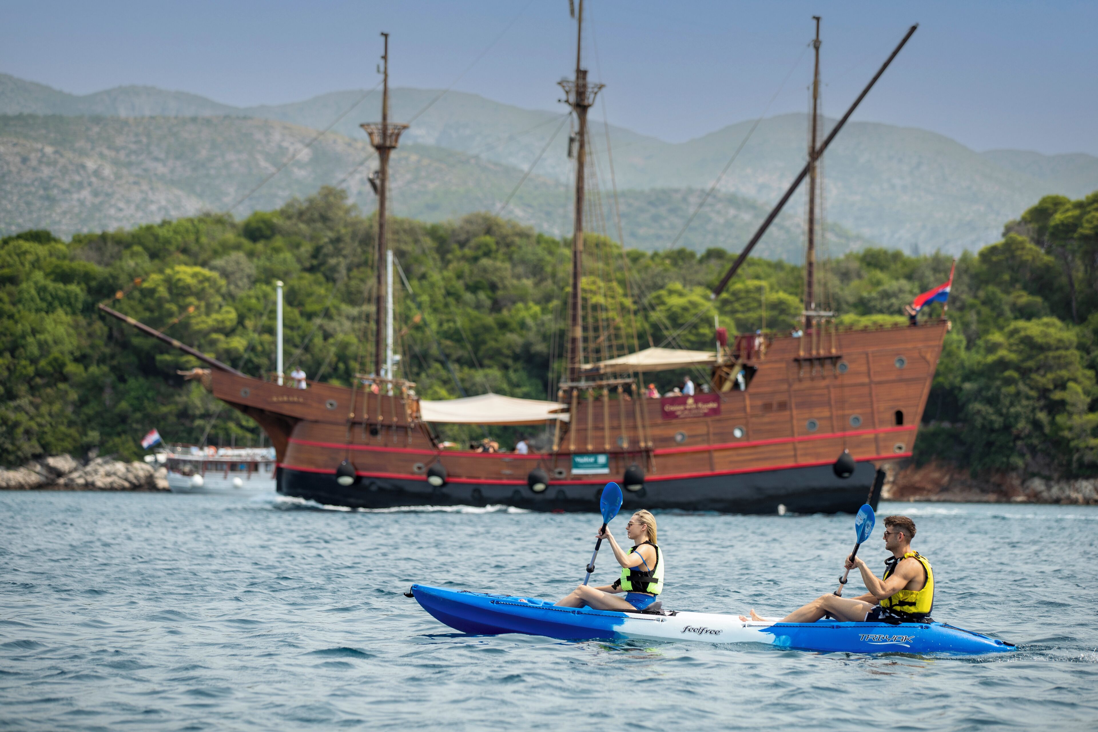 Two people kayaking on a calm body of water with a large ship in the background.