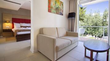 A hotel room with a bed, a sofa, a small table, a painting, and a view of trees through the window.