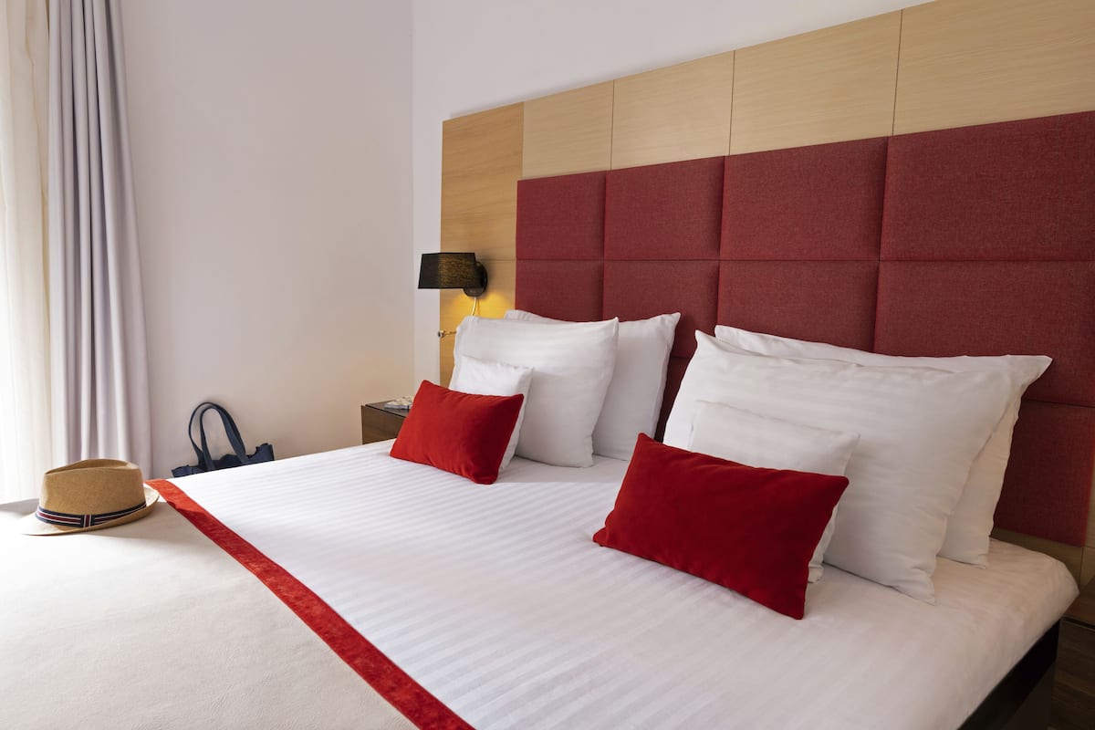 A hotel room with a large bed, red and white pillows, a wooden headboard, a nightstand with a lamp, and a hat on the bed.