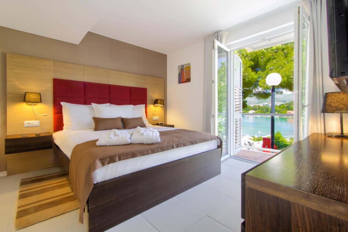 A modern hotel room with a large bed, a wooden headboard, and a view of the sea through a glass door.