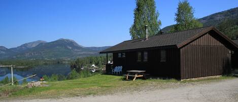 A cabin by a lake with a picnic table and chairs, surrounded by mountains.