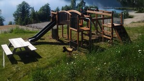 A playground with a slide and climbing structures near a lake with mountains in the background.