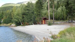 A cabin by a lake with a dirt path leading to it, surrounded by dense forest.