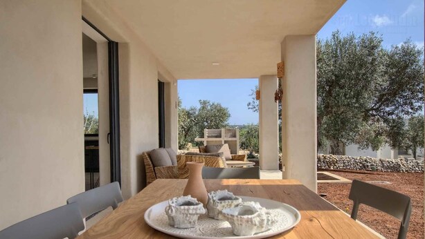 Una mesa de madera con un plato blanco de conchas marinas, un sofá de mimbre y vistas a olivos y un muro de piedra.