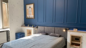 Un dormitorio con cama, mesita de noche, estante en la pared, un cuadro enmarco y un pequeño mueble de madera.