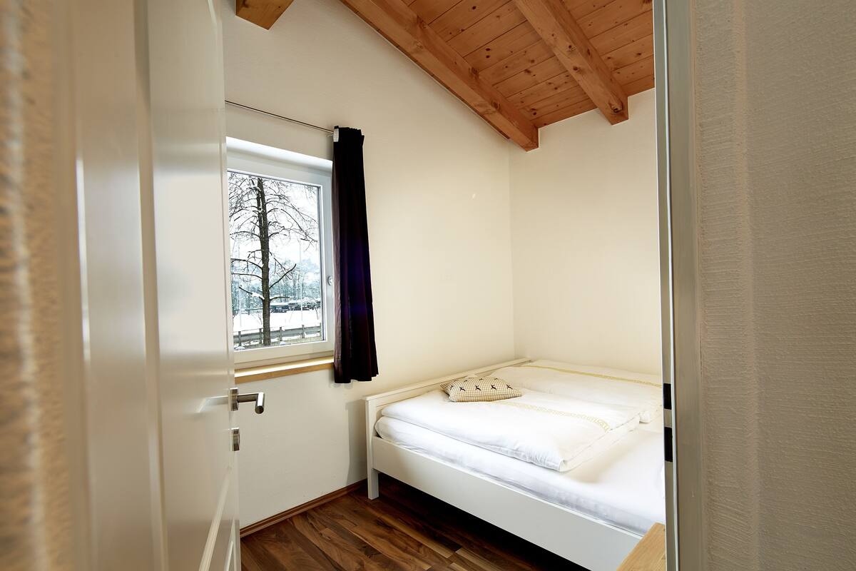 Una habitación pequeña con cama individual, techo de madera, ventana con vistas a árboles y puerta con manija.