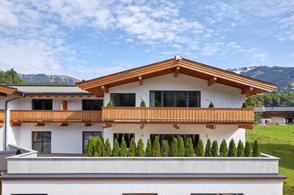 Una casa moderna de dos pisos con balcón de madera, rodeada de follaje y montañas al fondo.