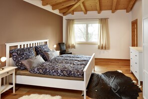 Un dormitorio con techo de madera, cama con ropa de cama estampada, mesita de noche, cómoda y una ventana grande con cortinas.