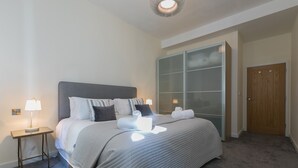 A neatly made bed with a grey headboard, a bedside table with a lamp, a large glass-fronted wardrobe, and a wooden door. - Sea Scapes – Sleeps 2/4 – Sea Views – Dog-Friendly - On-Site Parking (St Ives)