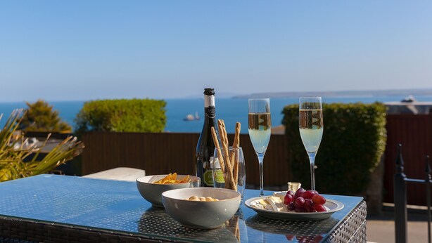 A table with a bottle, glasses, and food items, overlooking a scenic view. - Sea Scapes – Sleeps 2/4 – Sea Views – Dog-Friendly - On-Site Parking (St Ives)
