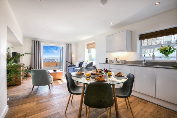 A modern kitchen with a dining area, a large window with a view, and a living room with a sofa and a TV. - Sea Scapes – Sleeps 2/4 – Sea Views – Dog-Friendly - On-Site Parking (St Ives)