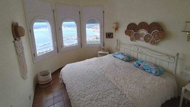 A bedroom with a bed, a ceiling fan, three windows with a view, a wall-mounted lamp, and a decorative headboard.