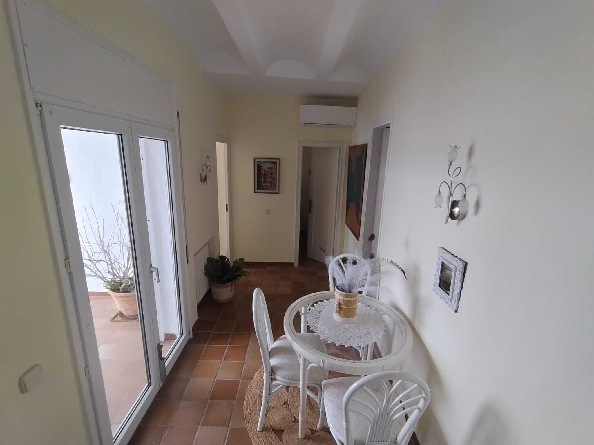 Un pasillo con una silla y una mesa blancos, una planta en maceta y una puerta que da al exterior.