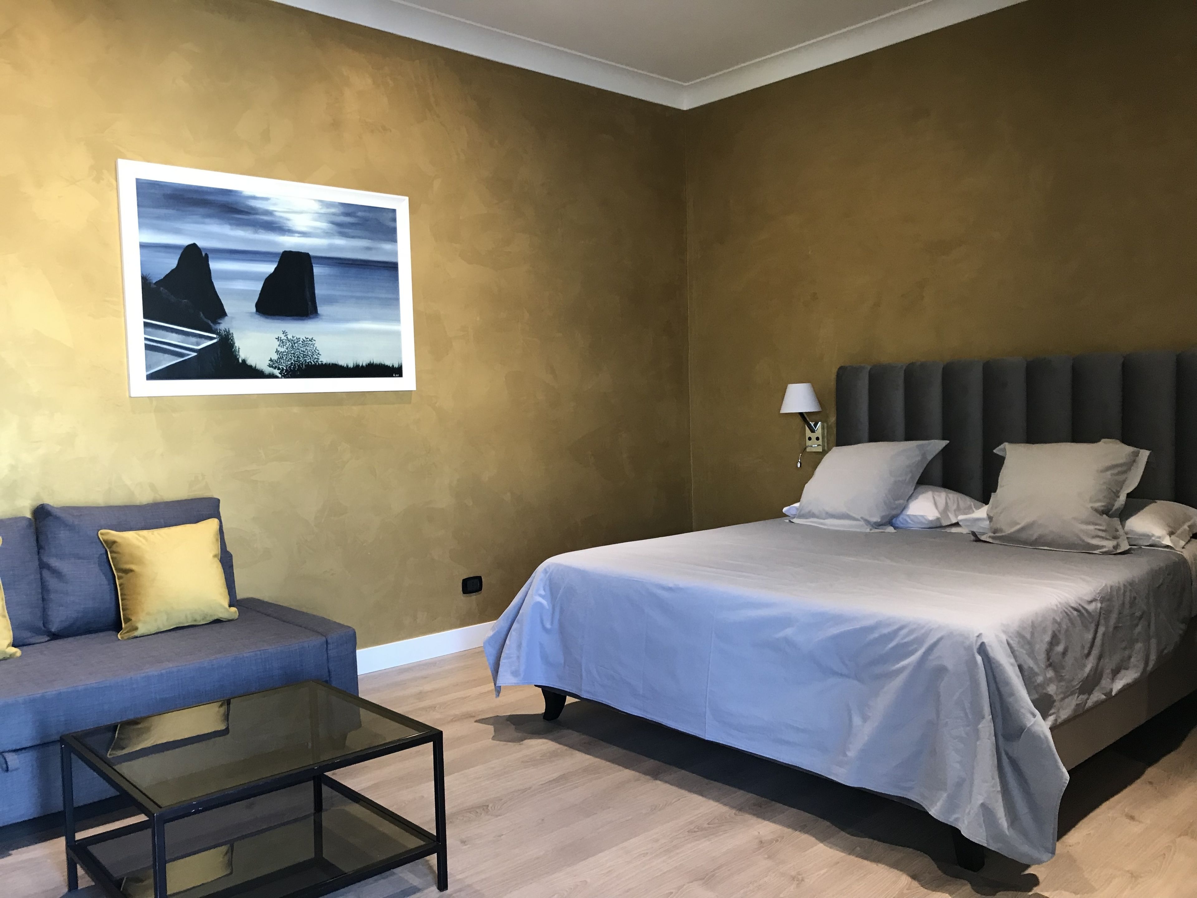 A bedroom with a bed, a sofa, a glass coffee table, and a wall-mounted lamp.