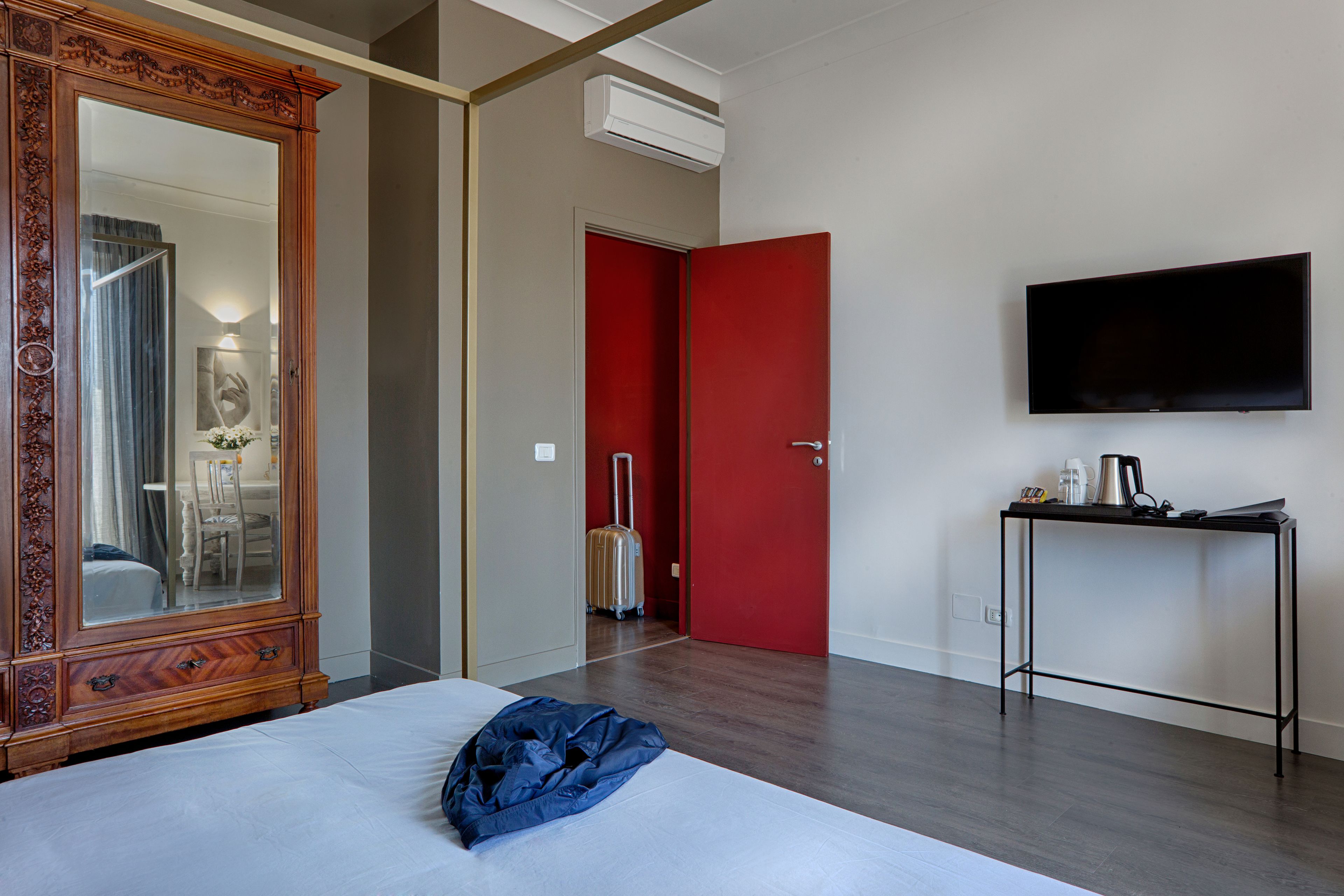 A hotel room with a large bed, a wooden wardrobe, a red door, a wall-mounted TV, and a small table with a kettle and cups.