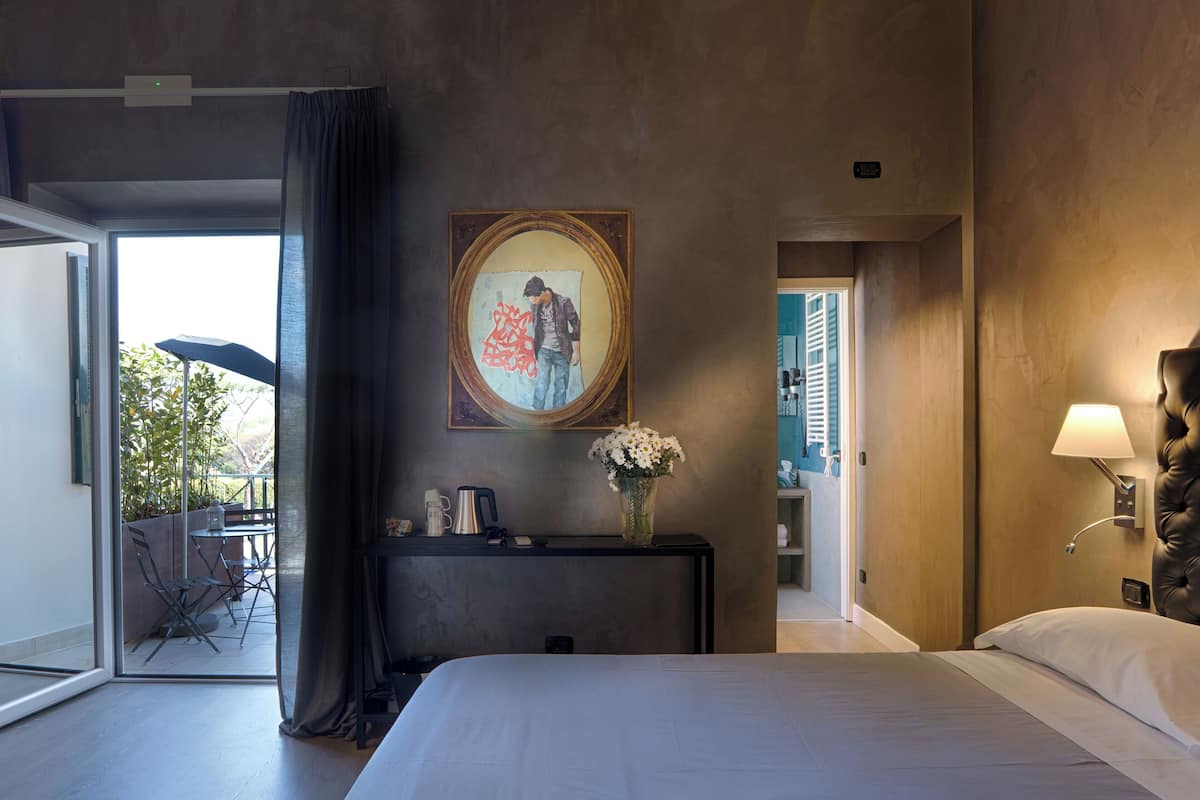 A hotel room with a bed, a desk with a vase of flowers, a painting, and a view of the outside.