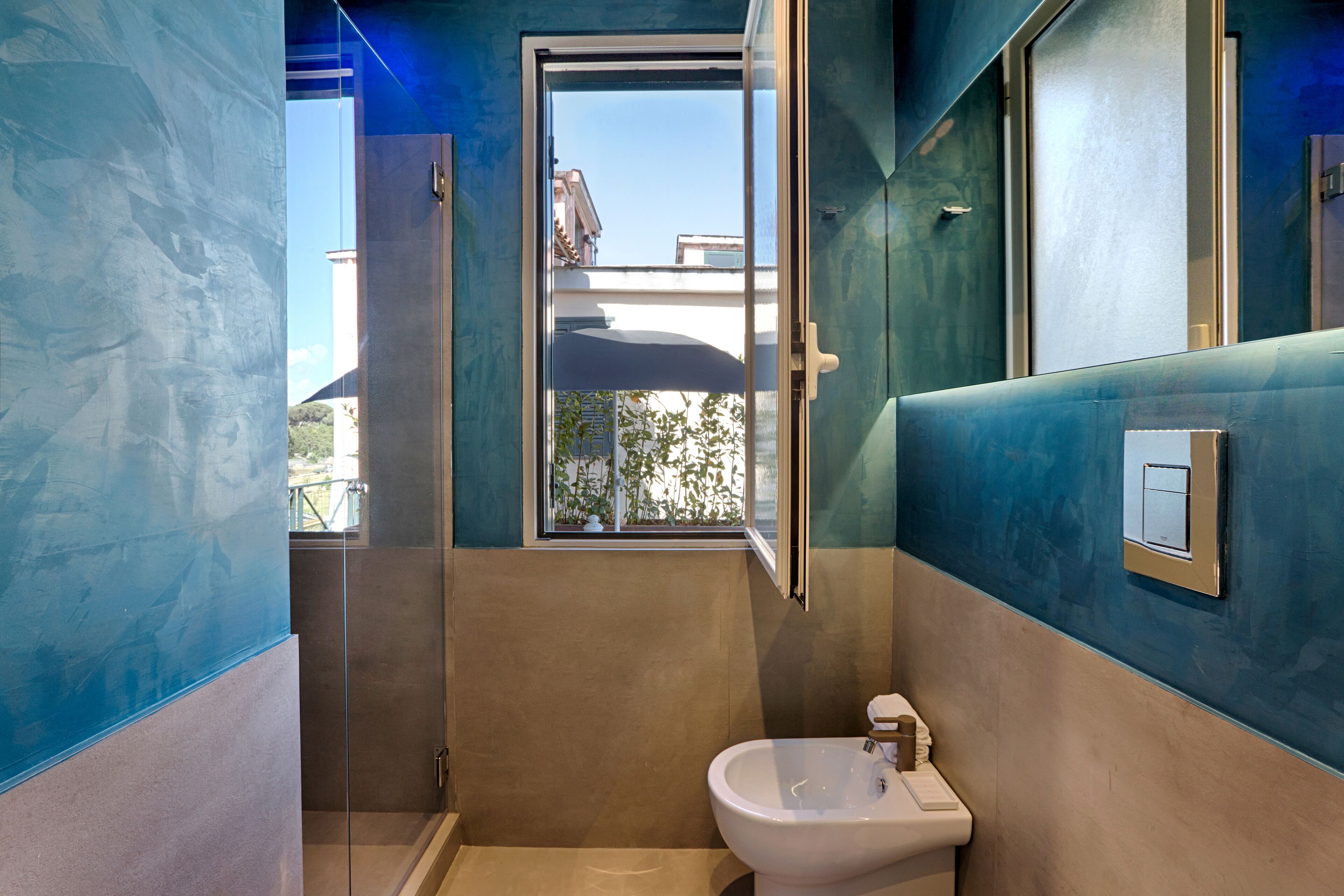 Un baño con cabina de ducha de vidrio, un lavabo blanco y una ventana con vistas a edificios y zonas verdes.