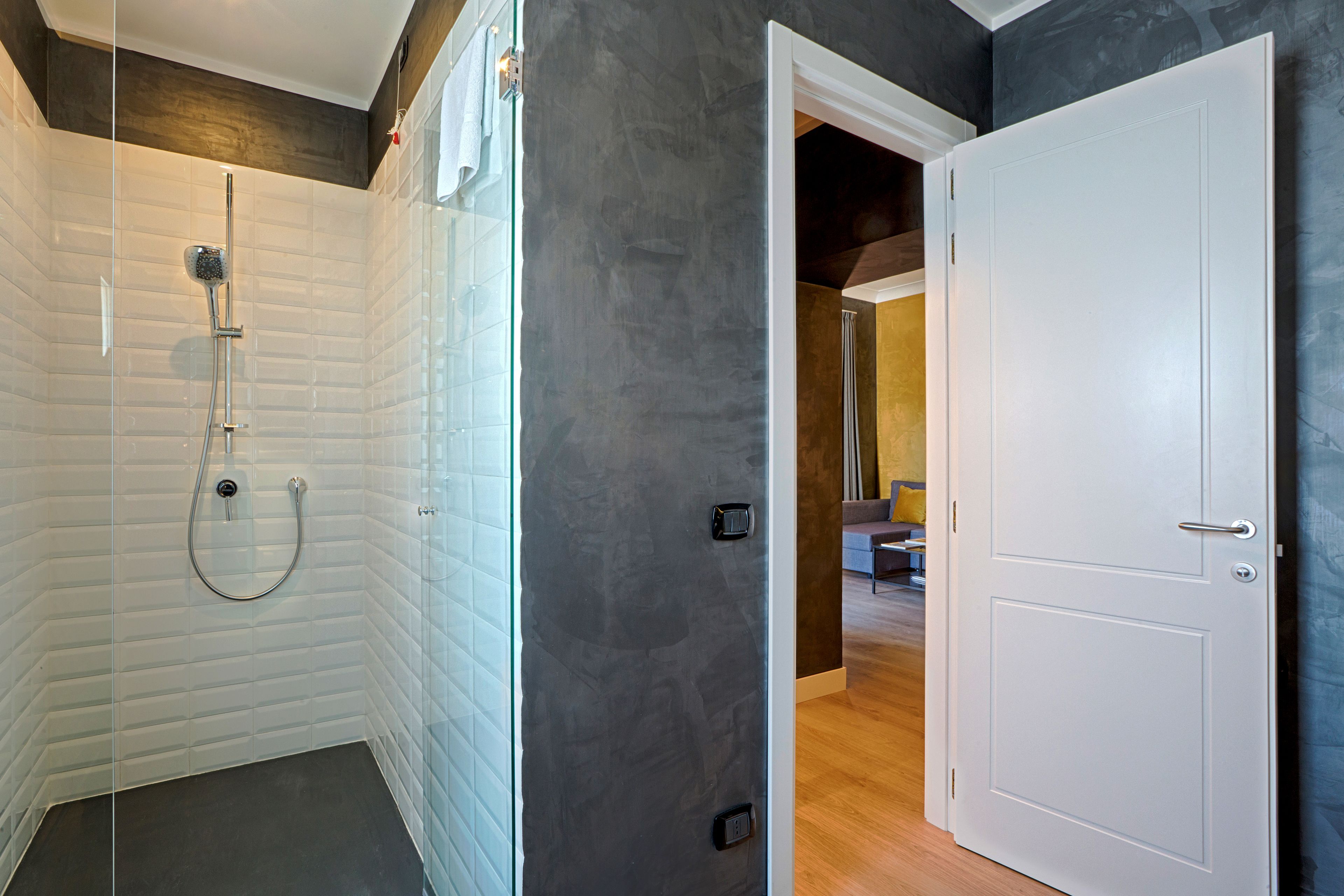 A modern bathroom with a glass-enclosed shower, a white door, and a visible hallway leading to a living area.