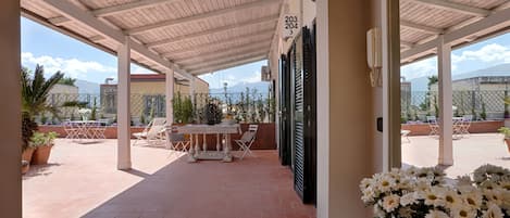 Uma área de varanda coberta com mesa, cadeiras, plantas em vasos e vista para a paisagem ao redor.