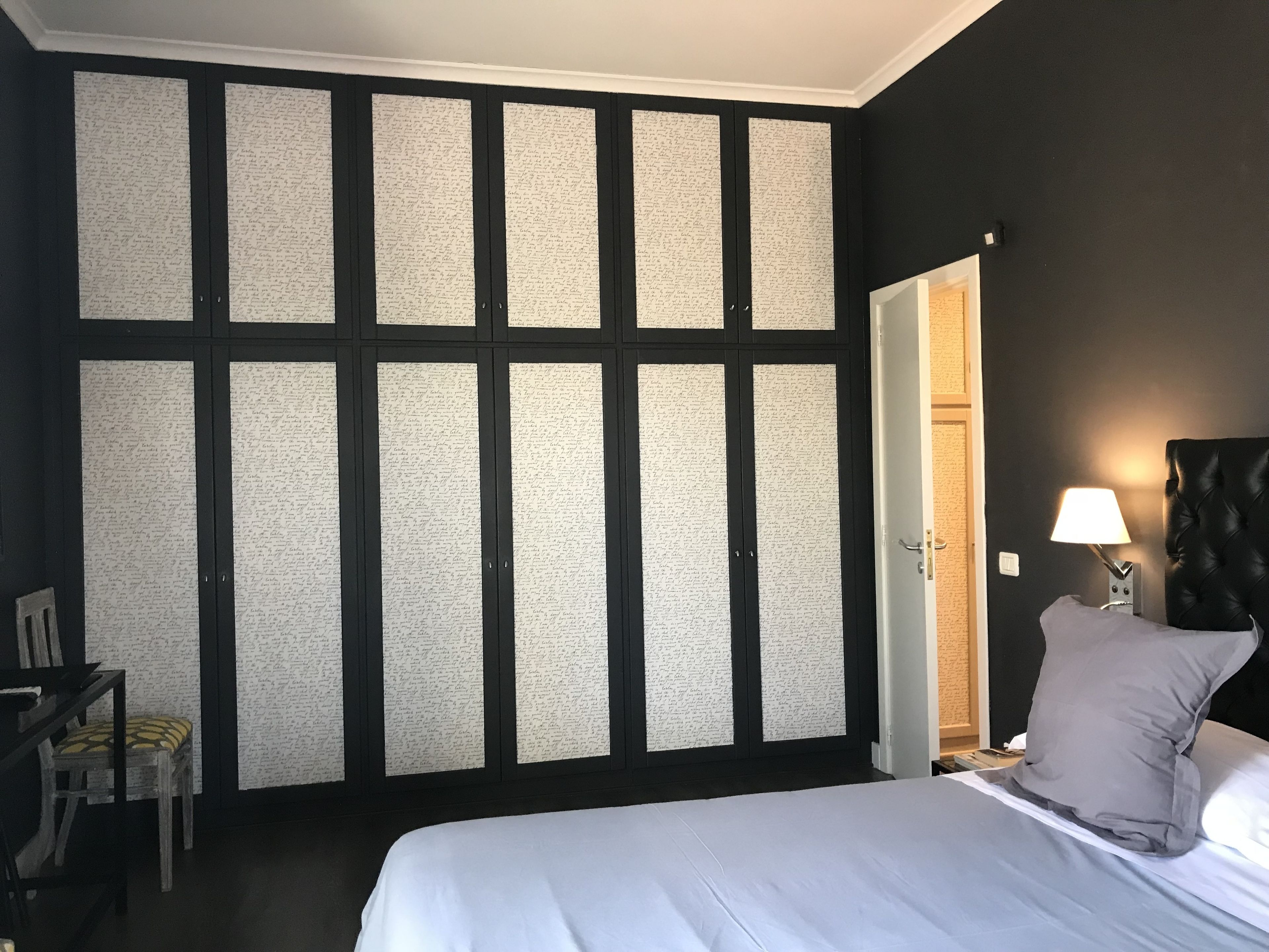 A bedroom with a bed, a nightstand with a lamp, a chair, and a wardrobe with a patterned back panel.