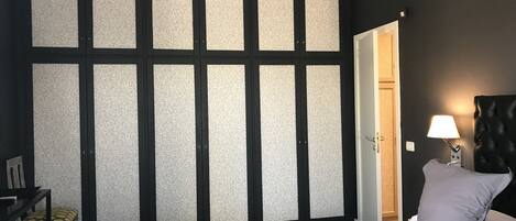 A bedroom with a bed, a nightstand with a lamp, a chair, and a wardrobe with a patterned back panel.