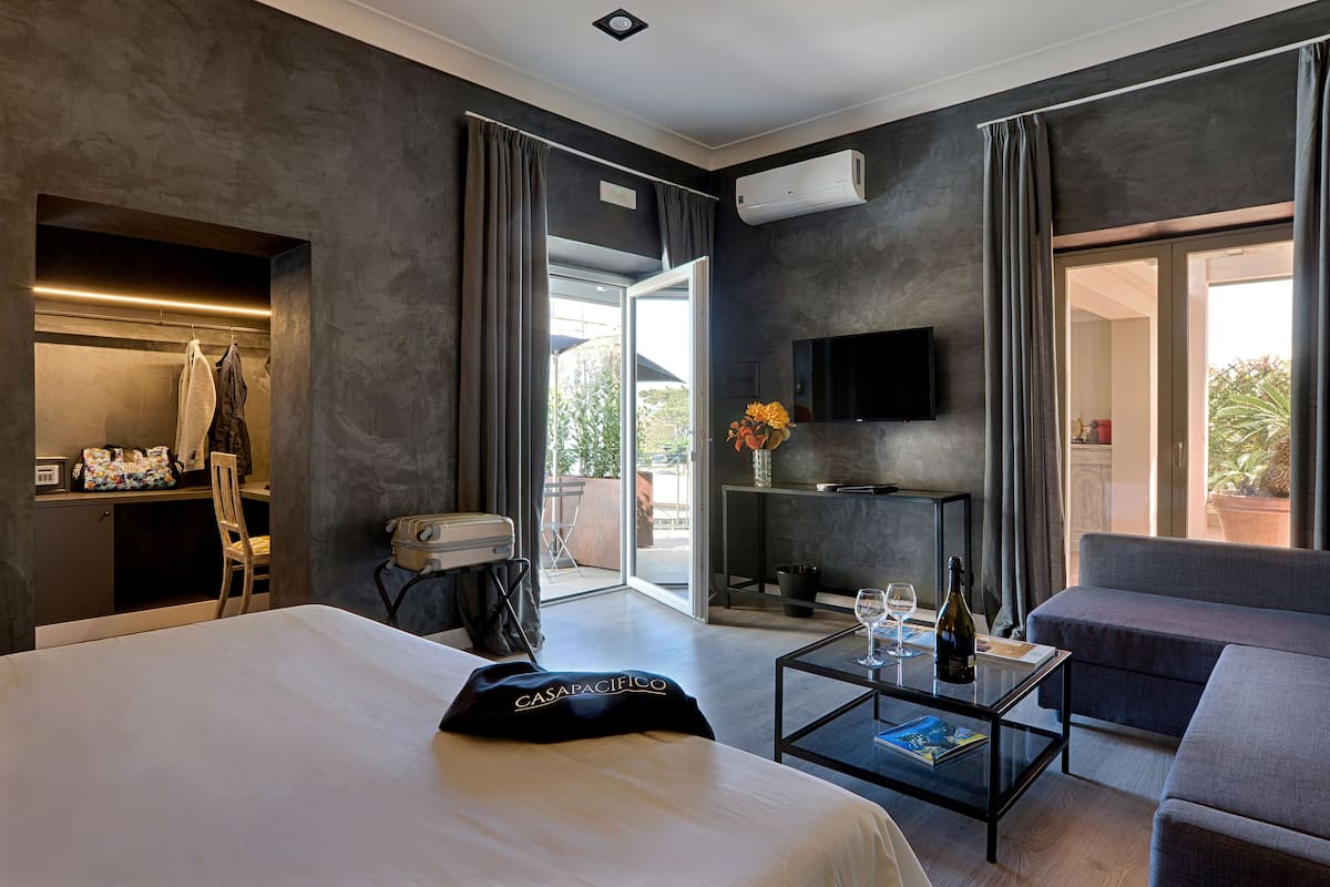 A modern hotel room with a large bed, a flat-screen TV, a coffee table with a wine bottle and glasses, a sofa, and a balcony with a view.