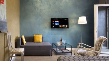 Ein modernes Wohnzimmer mit einem grauen Sofa, einem gemusterten Sessel, einem Glastisch und einem Wand-Fernseher, der einen Streaming-Dienst anzeigt.