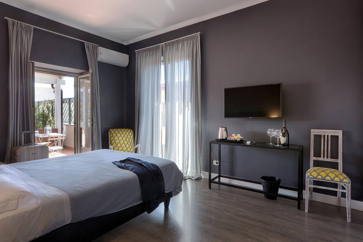 A hotel room with a large bed, a desk with a flat-screen TV, a chair, and a small table with a bottle and glasses.