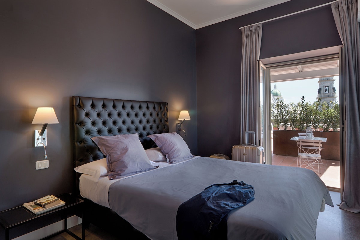 A hotel room with a large bed, two bedside lamps, a nightstand, a suitcase, and a view of a building through an open door.