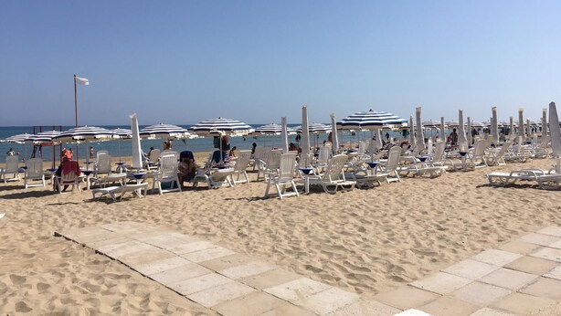 Een strand met rijen witte ligbedden en gestreepte parasols, een heldere lucht en de oceaan op de achtergrond.