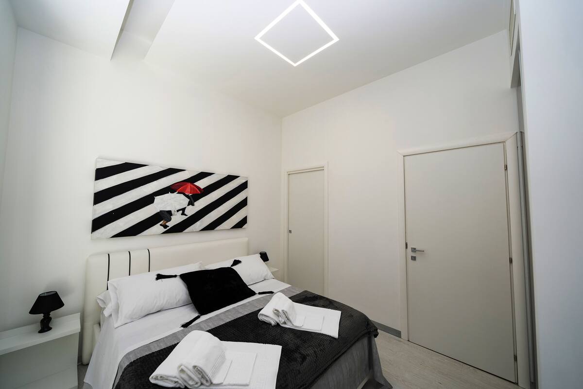 Ett modernt hotellrum med en snyggt bäddad säng, ett svart- och vitrandigt konstverk på väggen, en vit dörr och en taklampa.