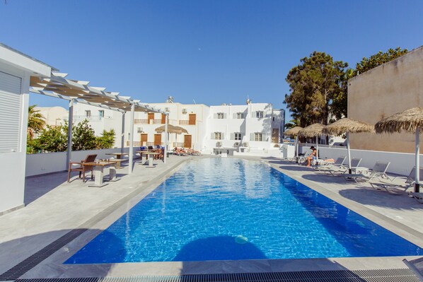 A swimming pool with lounge chairs and umbrellas, surrounded by white buildings with balconies.