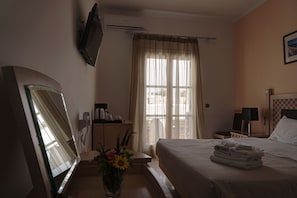 A hotel room with a bed, a desk with a telephone, a mirror, a TV mounted on the wall, a window with curtains, and a bedside table with towels.