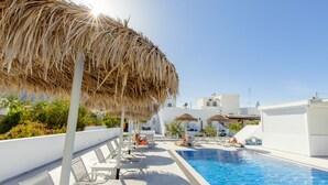 Zona de piscina en la azotea con sillones de descanso y sombrillas, rodeada de edificios blancos y follaje.