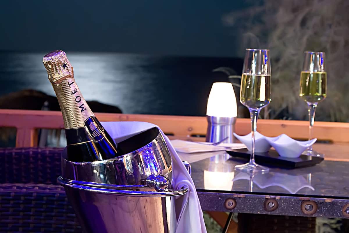 Uma garrafa de champanhe Moët, duas taças e uma vela acesa sobre uma mesa ao ar livre, com vista para o oceano e uma lua no céu.